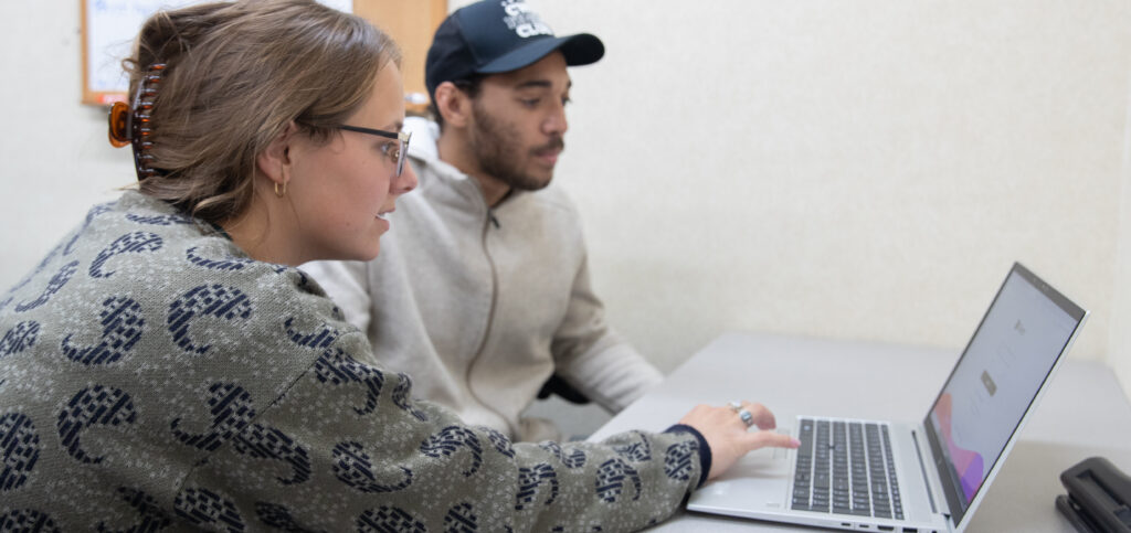 Two people sitting at a grey table, looking at a laptop