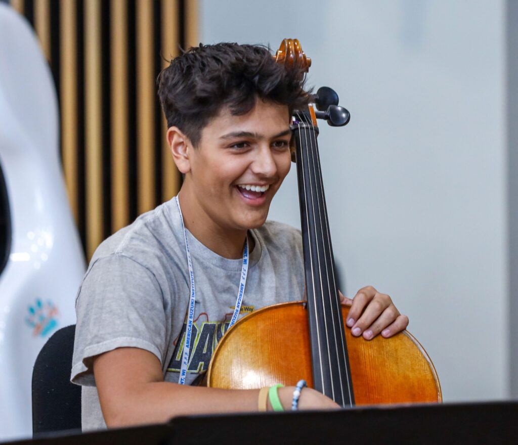 American Suzuki Institute teenage student smiling while playing cello in classroom.