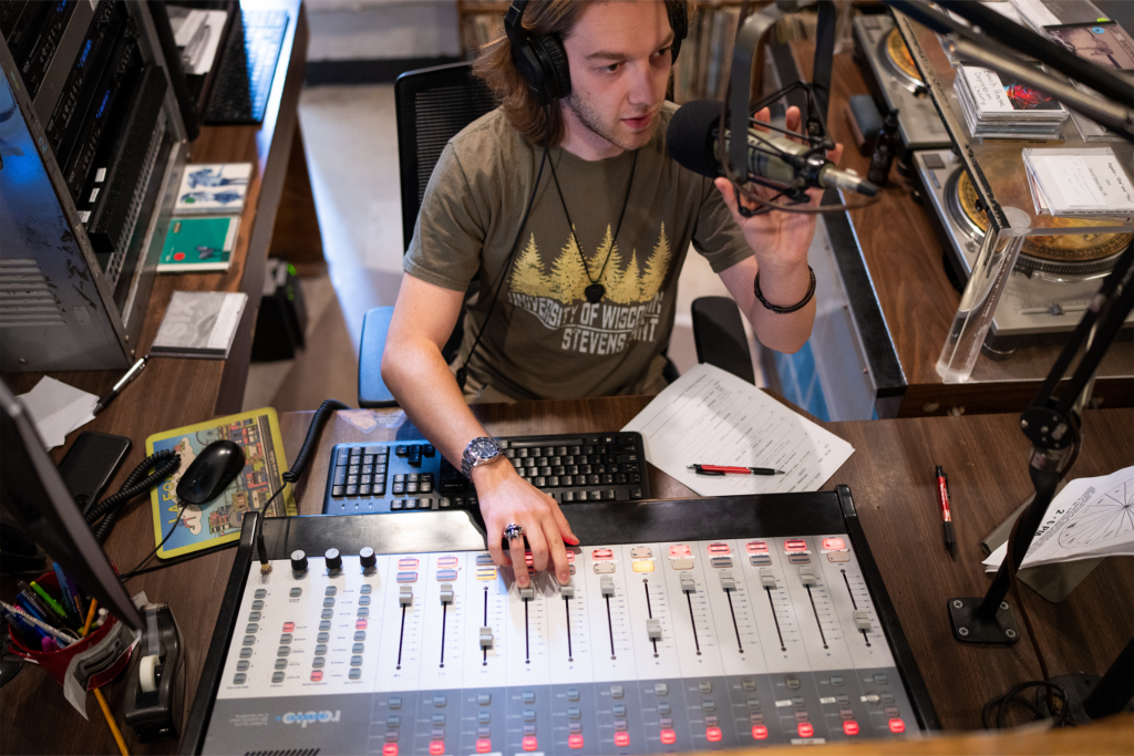 Student operates audio mixing board and microphone inside campus radio studio.