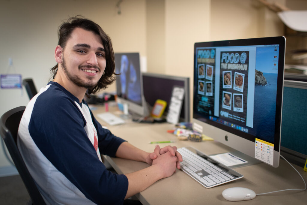 Student sits at computer workstation viewing food menu design project.