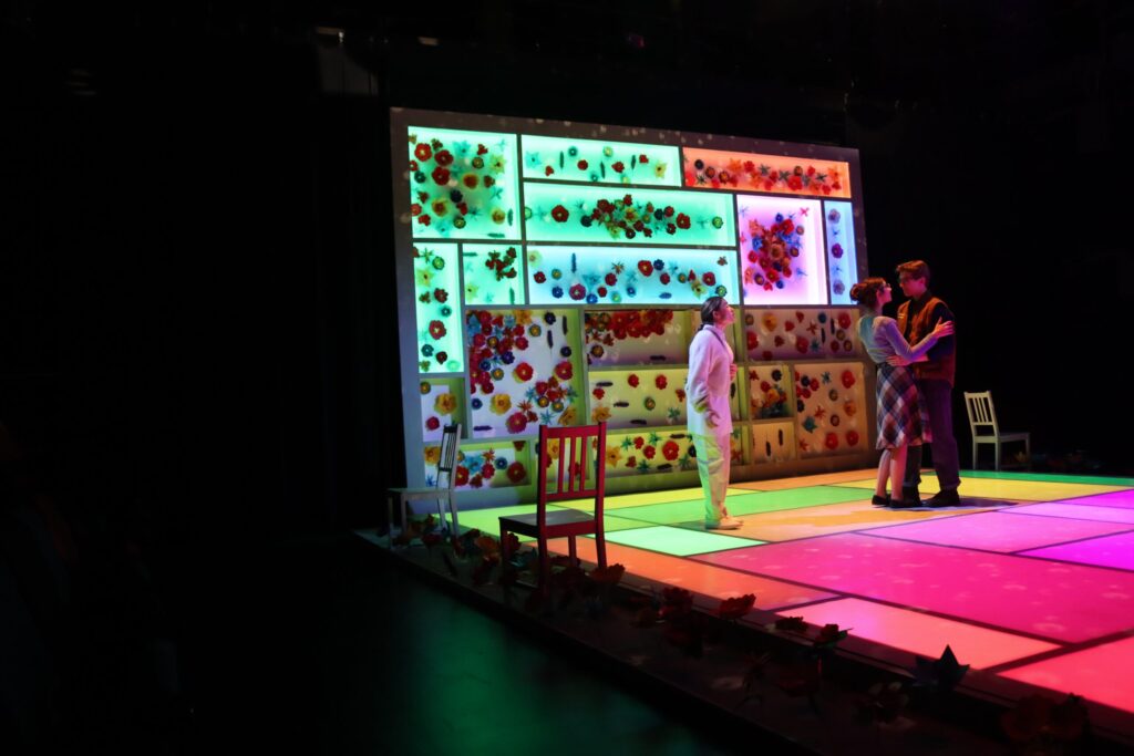 Three performers stand on colorful illuminated stage before vibrant floral backdrop.