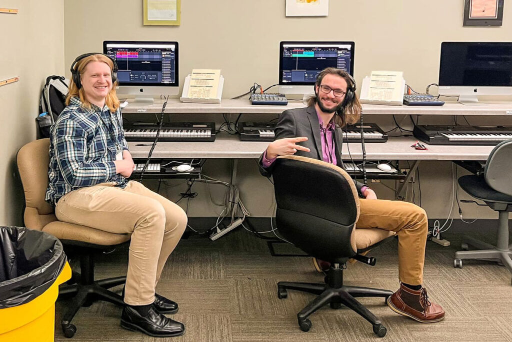 Two students work at music computer stations wearing headphones and chatting.