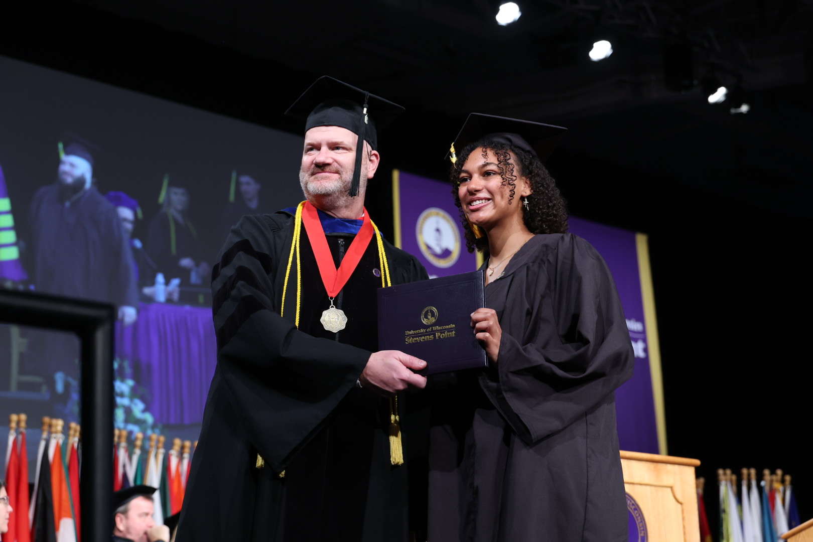 Dean Joshua Hagen stands near graduating student holding up UWSP diploma.