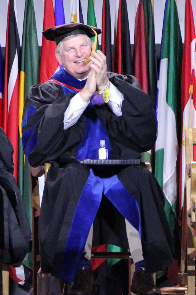 Faculty member in academic regalia applauding while seated in front of international flags.