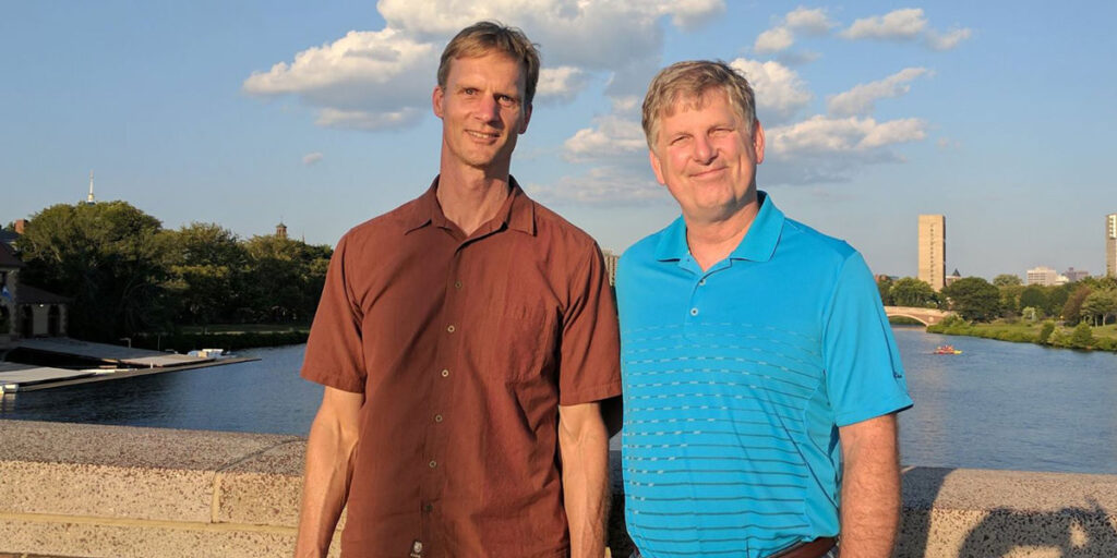 Two adults standing on a bridge with a river and city skyline behind.