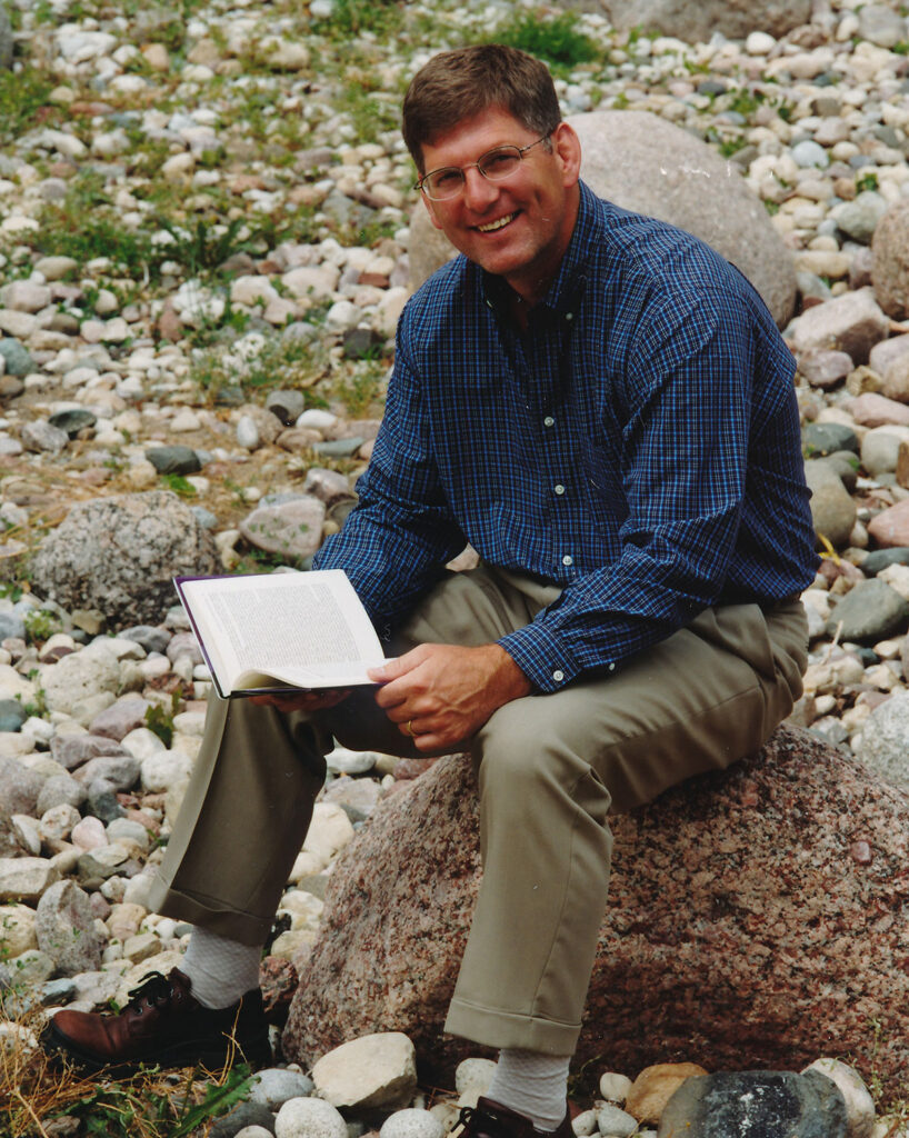Adult seated on a rock outdoors reading a book among stones and greenery.