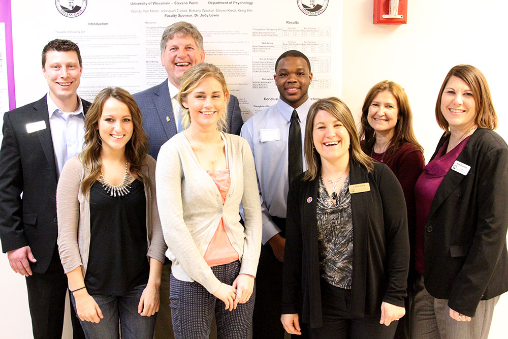 Presenters and attendees standing indoors in front of academic research posters.