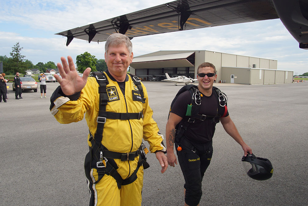 Two people wearing skydiving gear standing on an airport tarmac near small aircraft.