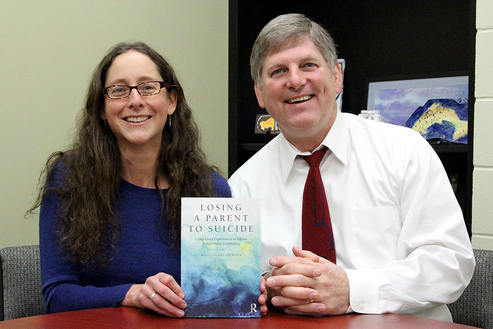 Two adults seated at a table holding a book titled ‘Losing a Parent to Suicide’.