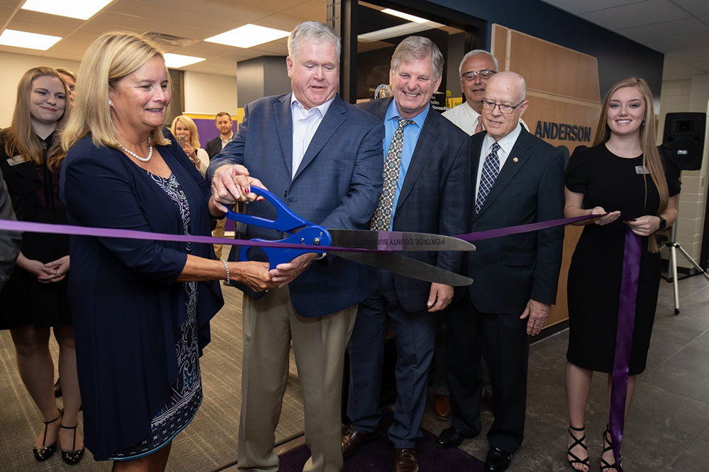 Ribbon-cutting ceremony indoors with participants holding oversized scissors.