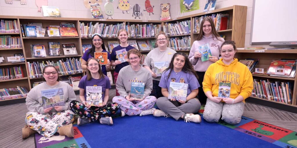 Group of preservice teachers holding books in an elementary school library.