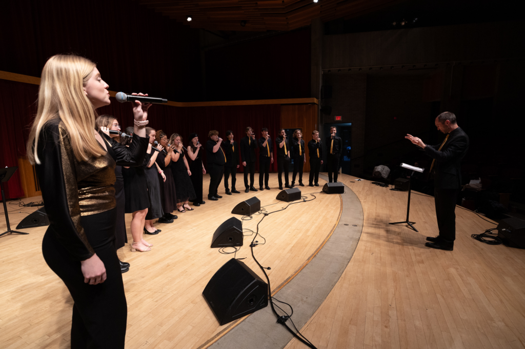Large vocal group performing onstage while conductor leads from front.
