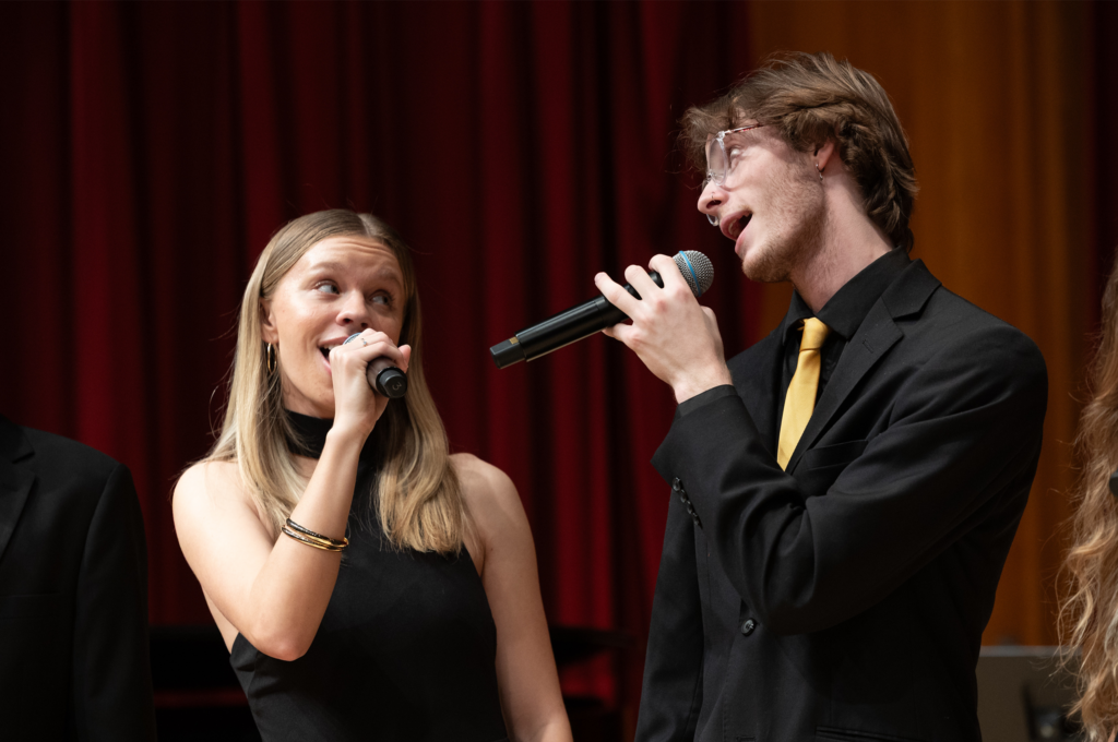 Two singers onstage performing with microphones against red curtain backdrop.