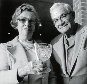 Historical black and white photo of an elderly man and woman with the woman holding a glass goblet