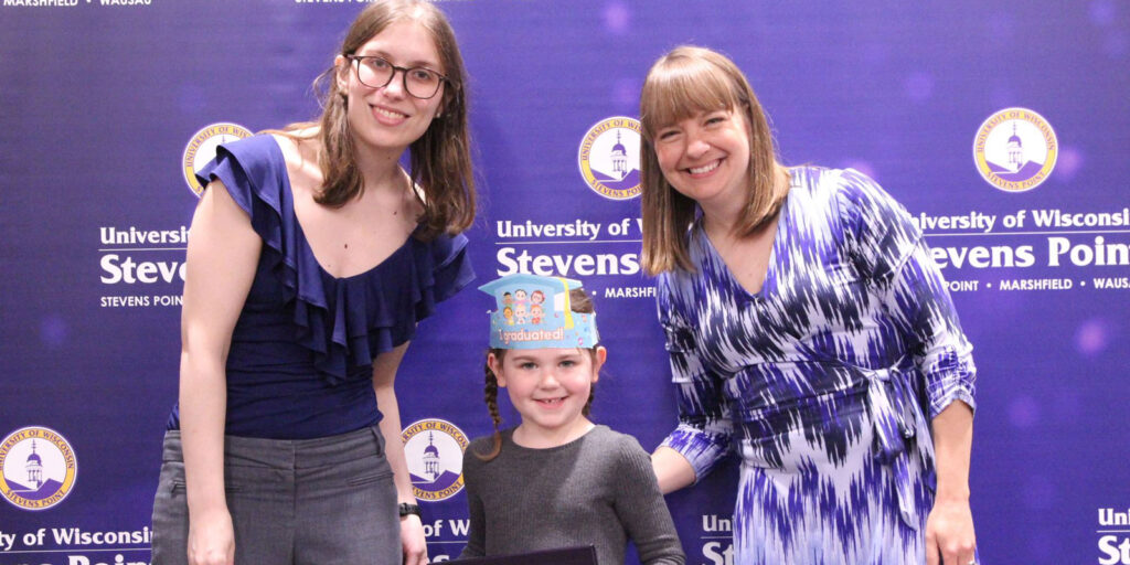 Child standing between two adults in front of a UW–Stevens Point backdrop at a recognition or celebration event.