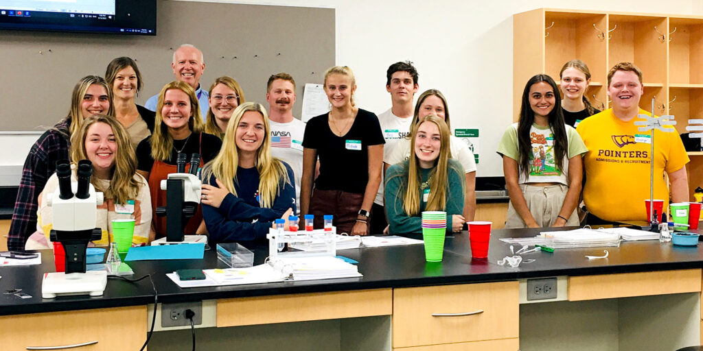 Large group of students gathered around lab tables conducting experiments using microscopes and plastic labware.