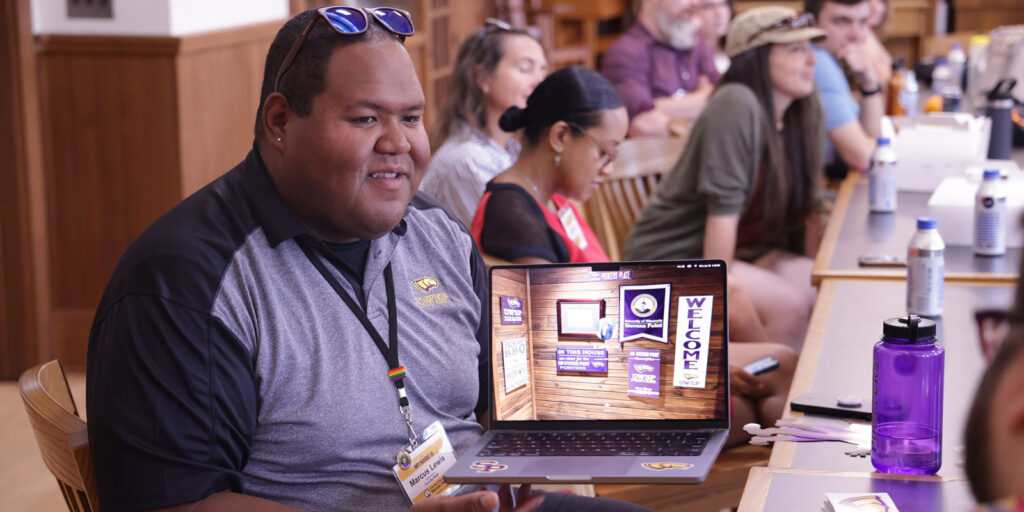 Instructor in a classroom holding a laptop open to a welcome screen for a digital orientation or program activity.