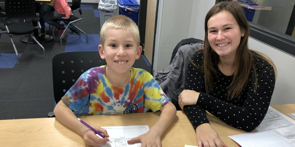 Child working on a worksheet at a classroom table with a student teacher seated beside them providing support.