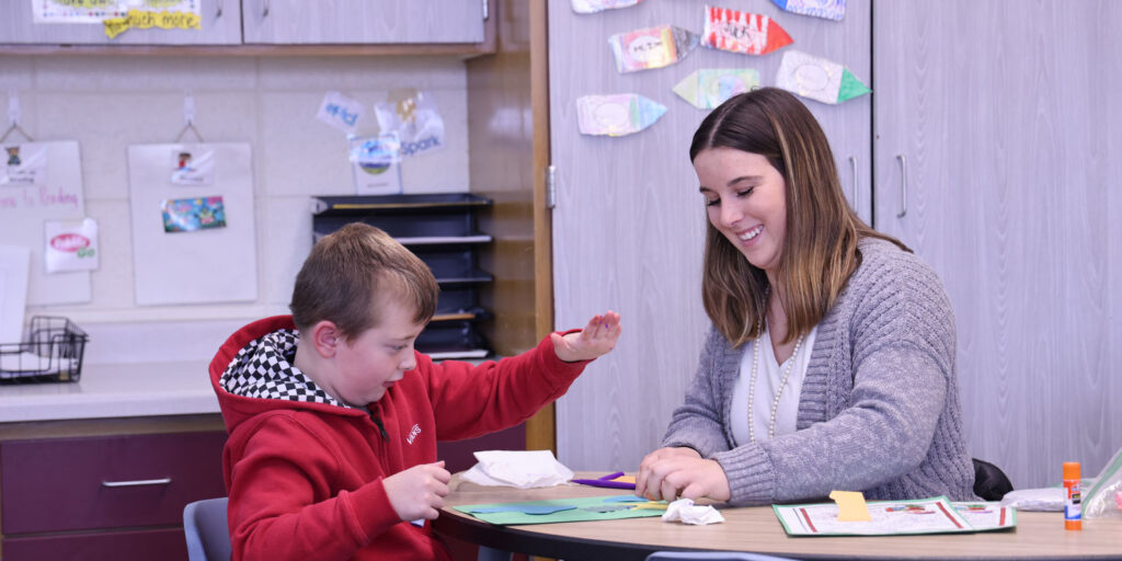 Student teacher working at a small table with a student during a hands-on craft activity using glue and paper.