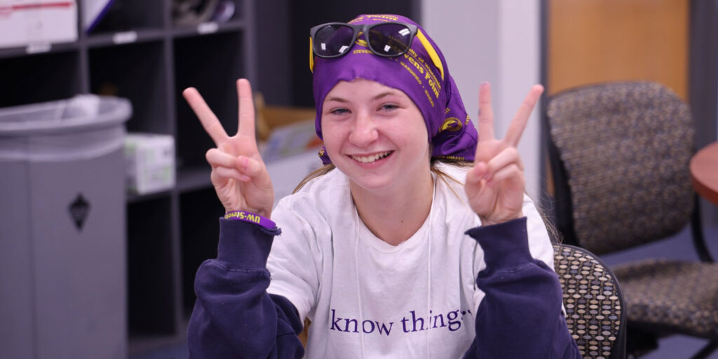 A student wearing a program T-shirt flashes peace signs while seated in a classroom.