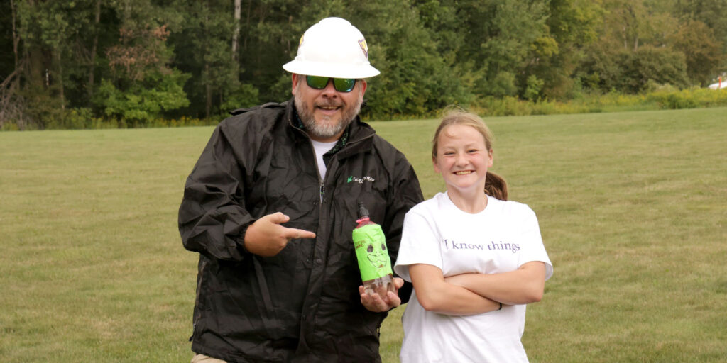 A person wearing a hard hat and work jacket stands next to another person holding a decorated plastic bottle outdoors in a grassy area.