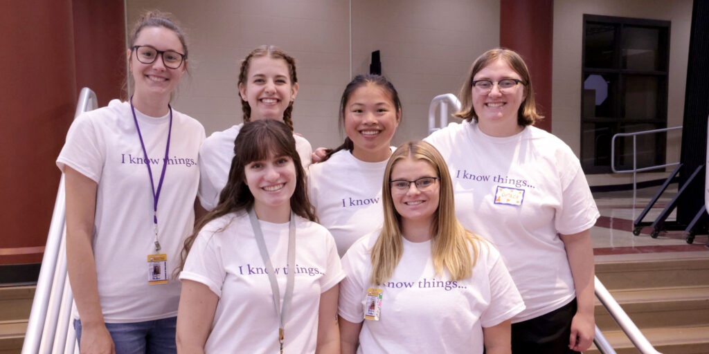 A group of people wearing matching “I know things…” shirts stand together inside a building near a stairway.