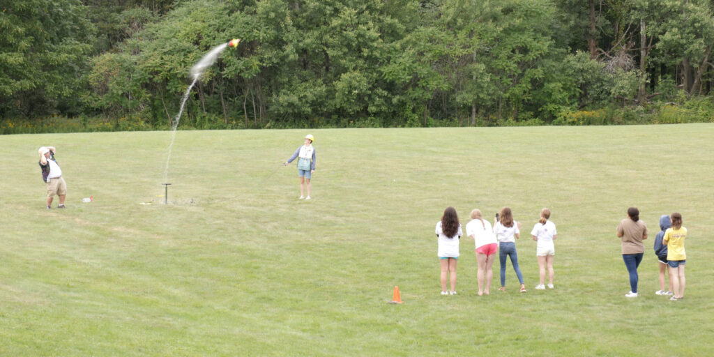 A group of students on a grassy field watch as a bottle rocket launches into the air during an outdoor activity.