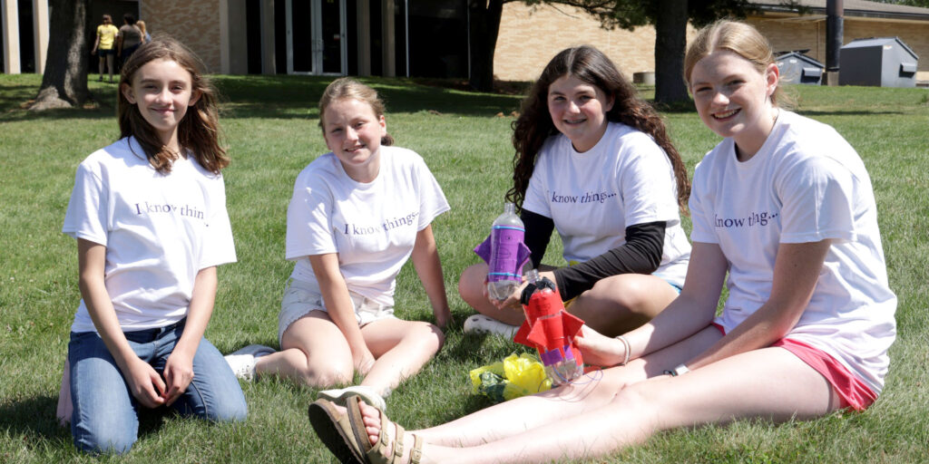 Four children sit on the grass outside holding decorated plastic rockets during an outdoor activity.