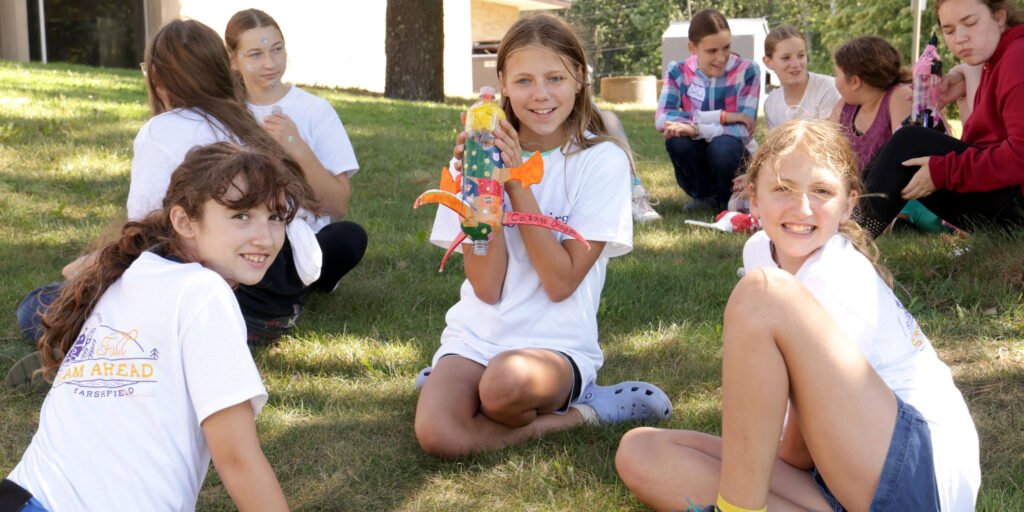Children sit on the grass holding colorful handmade craft rockets during an outdoor program activity.