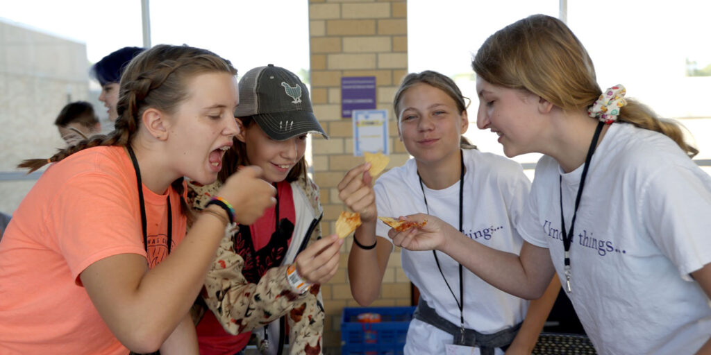 A group of students stand in a circle holding pieces of food during a shared activity in a bright indoor space.