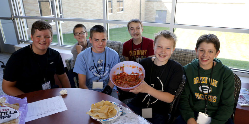 A group of students sit together at a table holding a large bowl of homemade salsa with chips on a plate nearby.