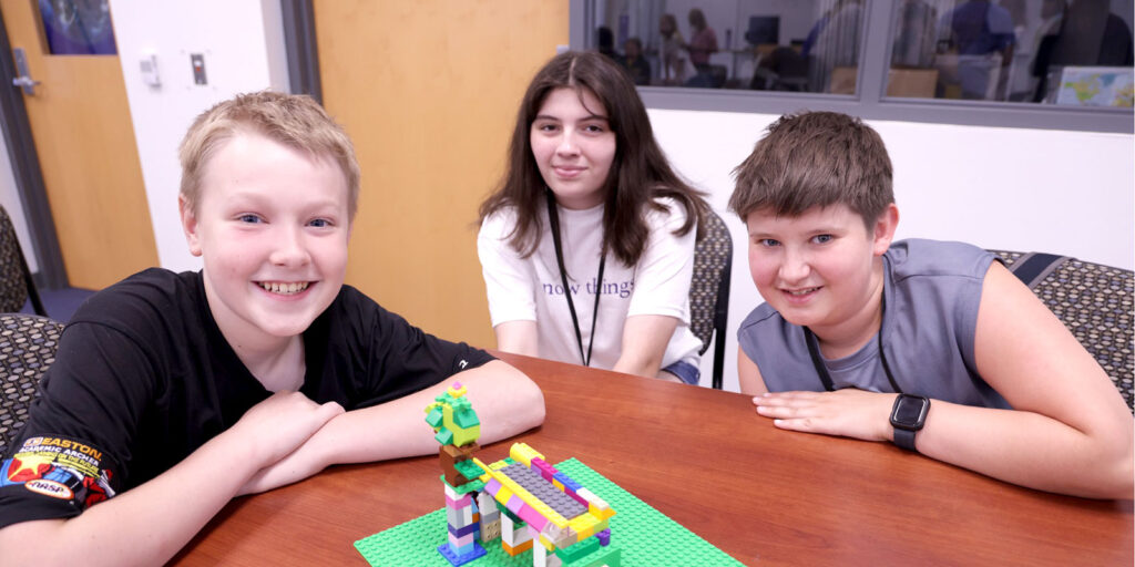 Three students sit at a table displaying a small LEGO project built on a green baseplate.