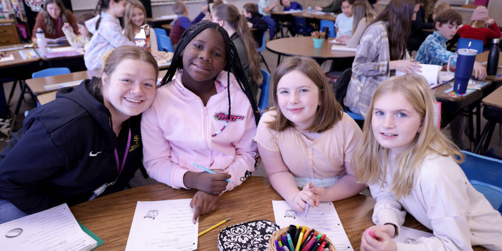 A group of students sit together at a classroom table working on worksheets during a literacy activity.