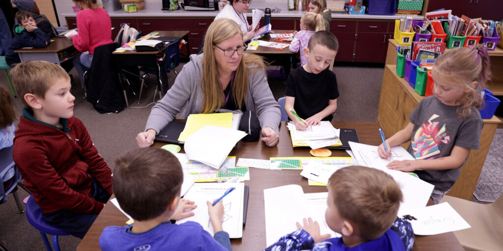 An educator meets with a small group of students at a classroom table while they complete writing activities.