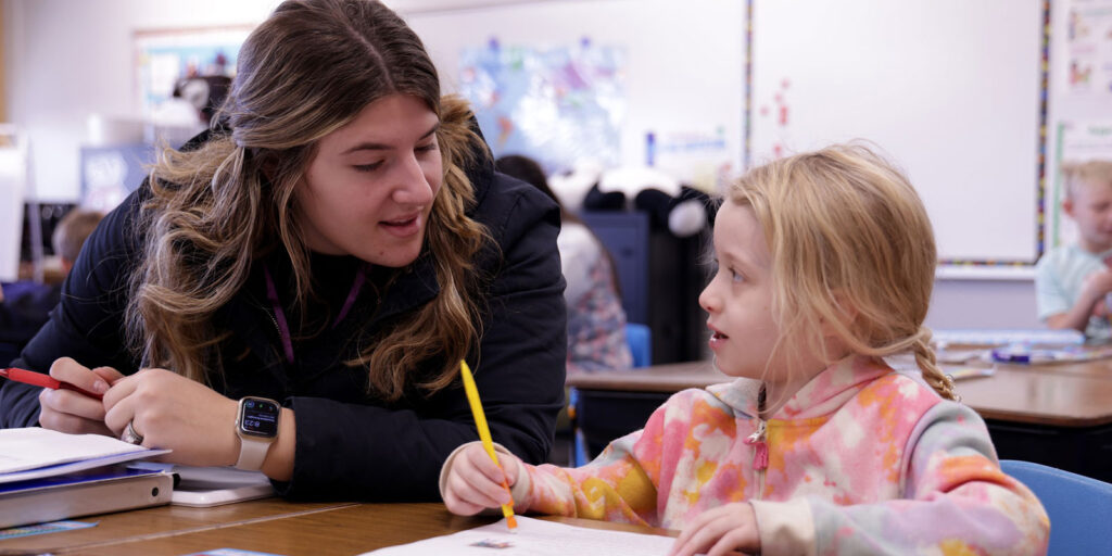 An educator sits beside a student who is writing in a notebook during a literacy exercise.