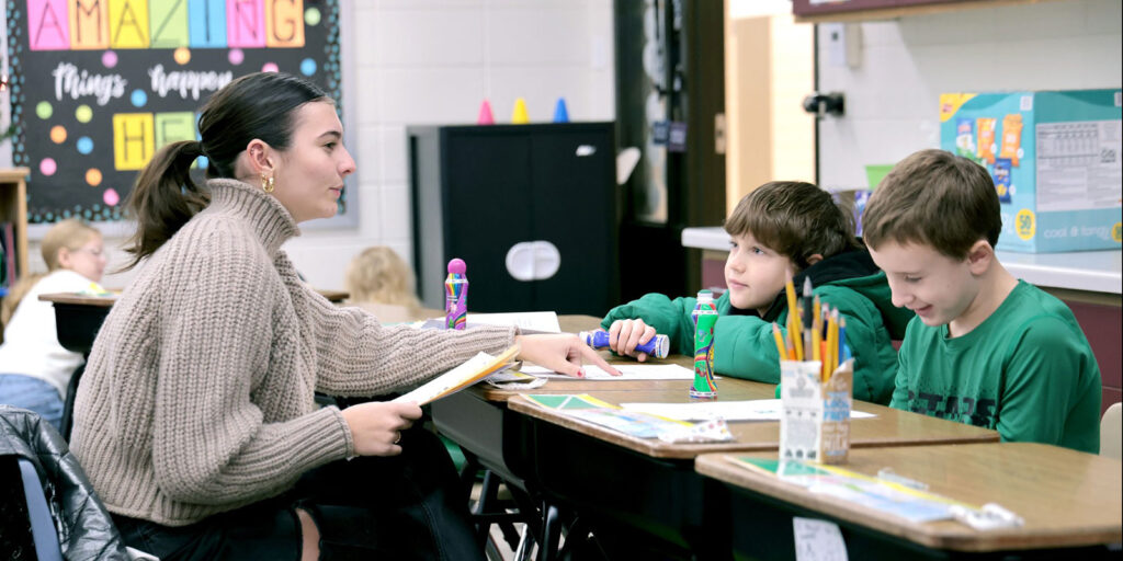 An educator works with two students at a table using colorful manipulatives during a classroom activity.
