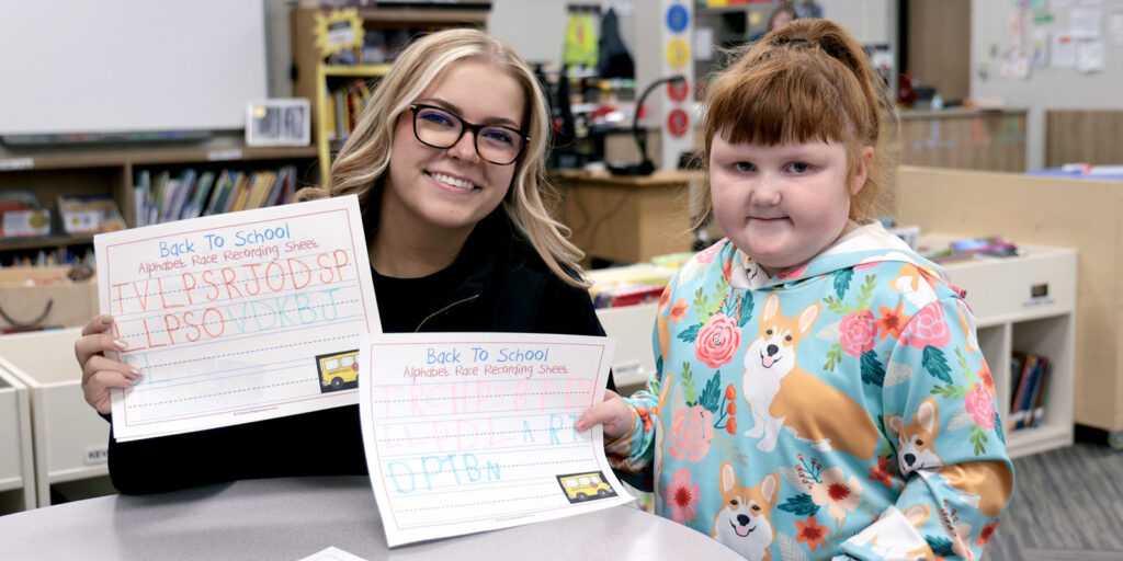 A student completes a handwriting and spelling exercise at a classroom desk.