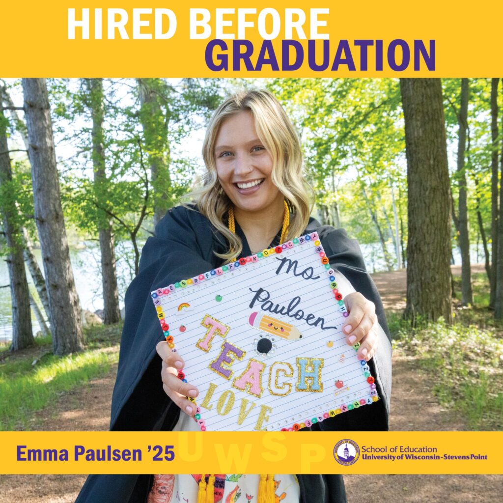 Student outdoors in a wooded lakeside area holding a decorated graduation cap with “Teach Love” written on it and “Hired Before Graduation” at the top.