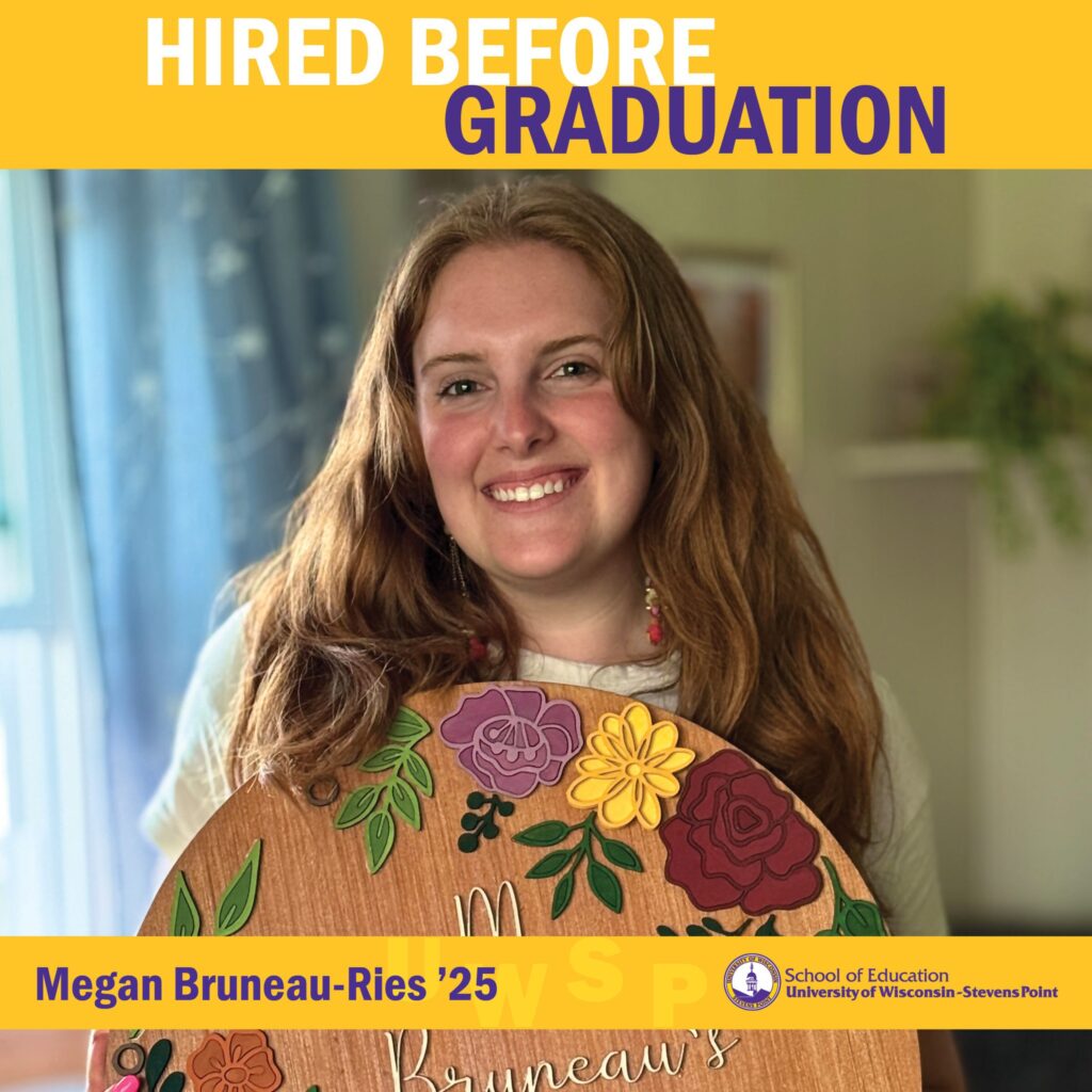 Student indoors holding a round wooden sign decorated with painted flowers under “Hired Before Graduation.”