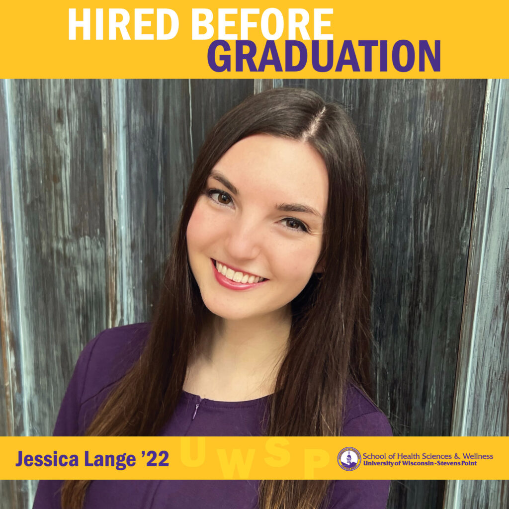 A graduate wearing a purple shirt stands in front of a weathered wooden backdrop with a “Hired Before Graduation” banner.