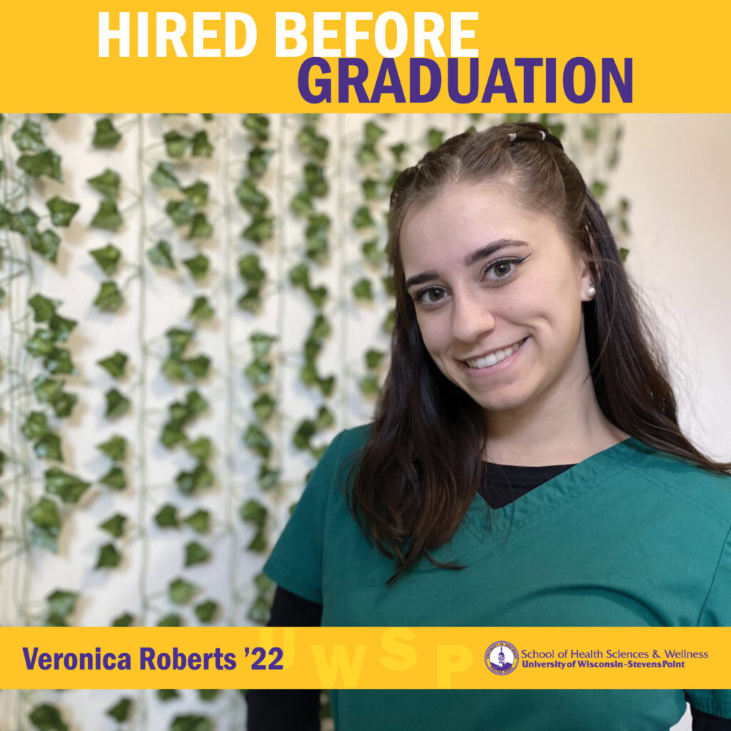 Graduate in scrubs poses in front of a wall decorated with hanging greenery under a “Hired Before Graduation” banner.