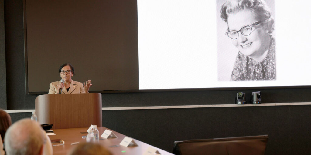 Chancellor speaking at a podium with an archival photo displayed on the screen.
