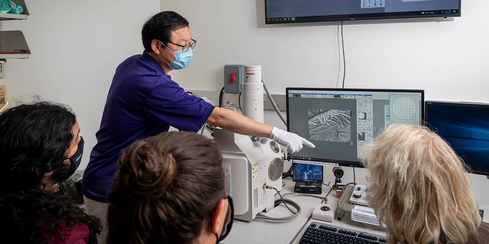 Professor and students looking at biology microscope and computer screen.