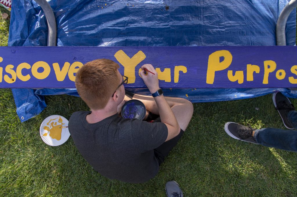 Student painting the phrase "discover your purpose" on a picnic table.