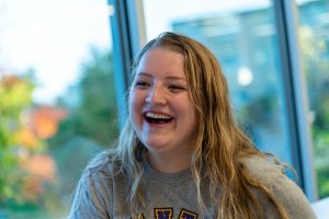 A female student laughing with a window view of fall on campus.