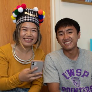 A male and female student smiling at the camera while the female holder her phone.