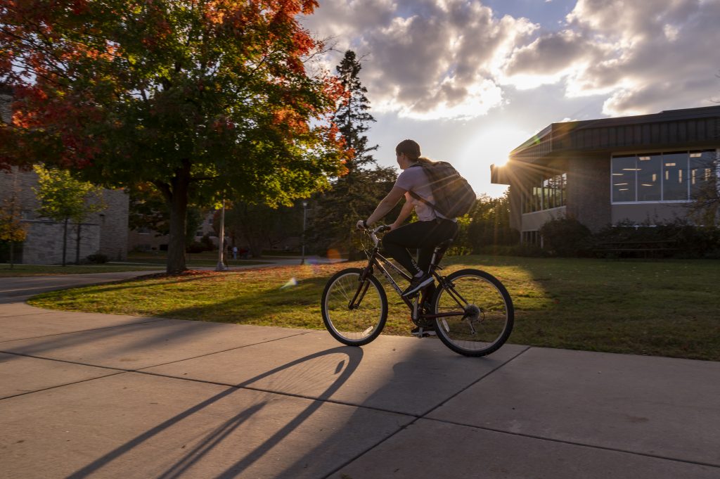 Student riding a bike on the sidewalk by the DeBot Dining Center at sunset.