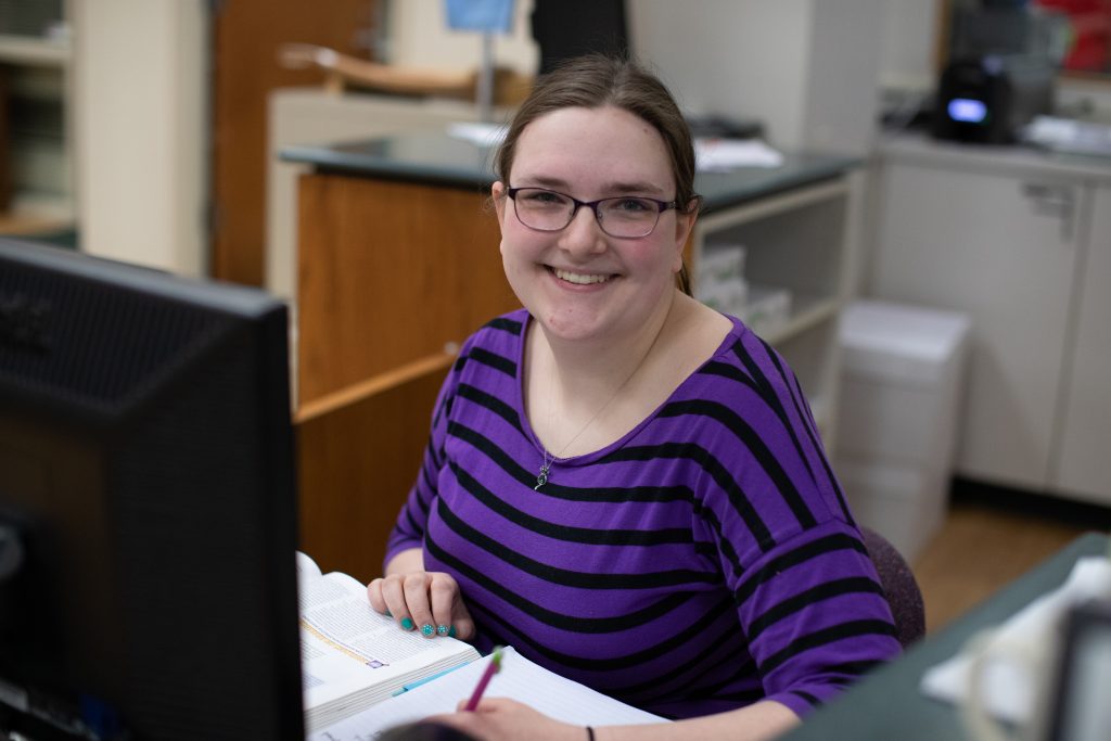 Student in a purple shirt smiling and studying at a computer.