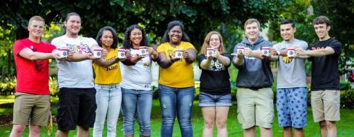 Five male and four females posing in a straight line outside holding Greek life coffee mugs.