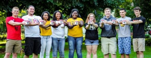 Five male and four females posing in a straight line outside holding Greek life coffee mugs.